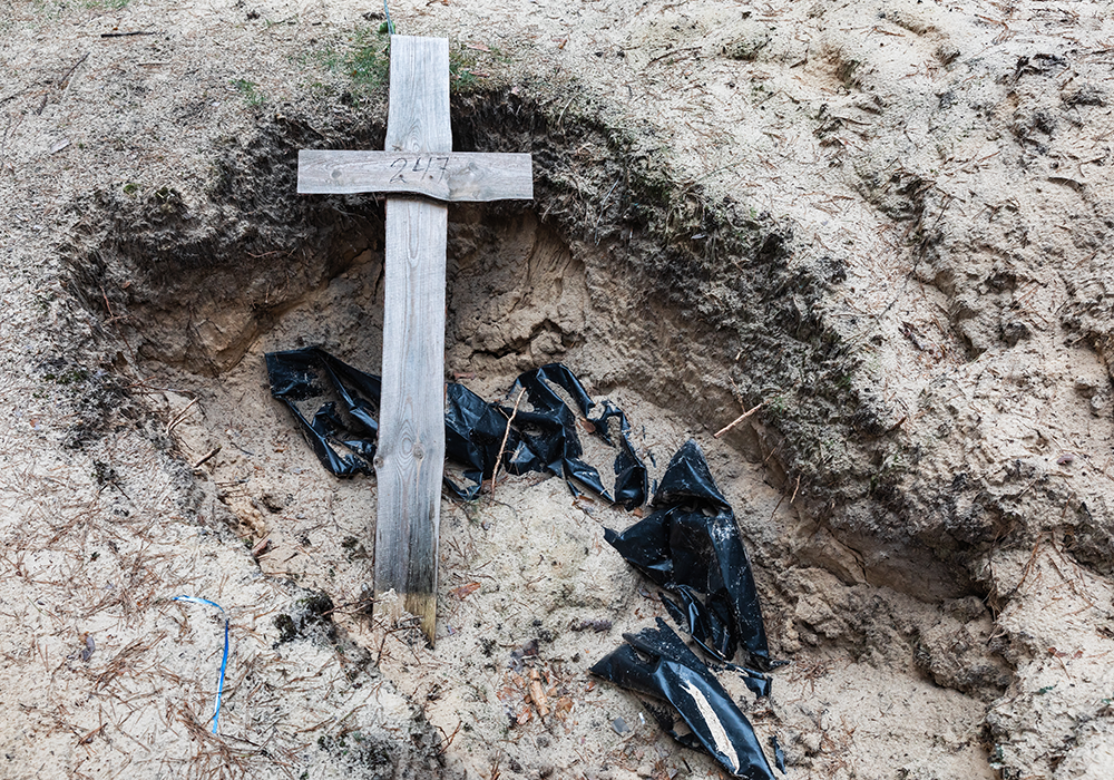 Une croix latine en bois est posée sur la terre. On devine la présence d’un corps dans une bâche noire sous la croix.