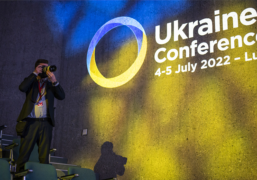 A photographer takes pictures on a stage with the logo of the Ukraine Recovery Conference of July 4 and 5, 2022 in the background.